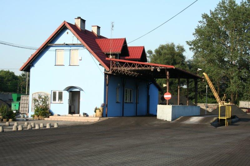 Niebieski budynek z czerwonym dachem, prawdopodobnie biuro lub magazyn, z rampą załadunkową i niewielkim dźwigiem na zewnątrz, teren wyłożony kostką brukową pokrytą pyłem.