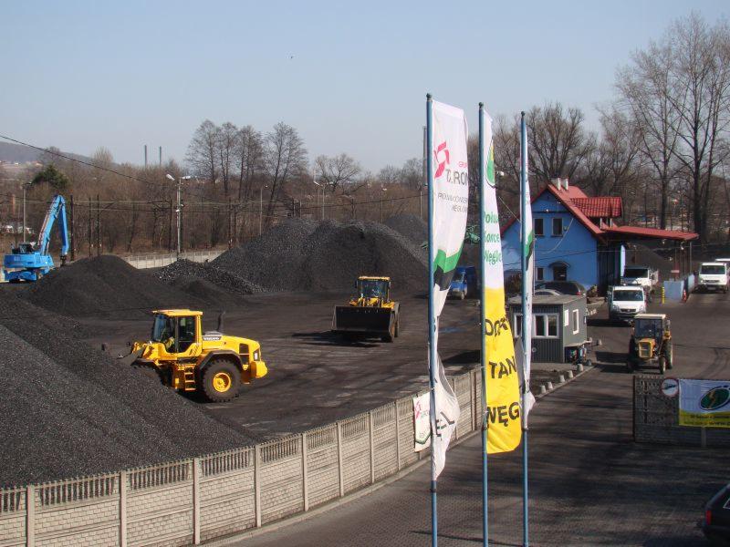 Żółta ładowarka Volvo na placu z hałdami czarnego materiału, w tle budynek biurowy i reklamy na masztach flagowych.