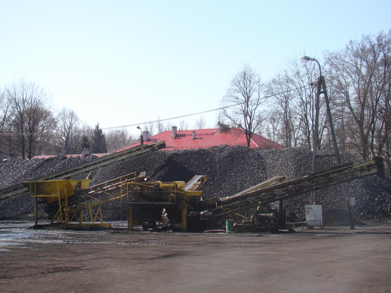 Kopalnia odkrywkowa z taśmociągiem transportującym czarny materiał sypki, w tle budynek z czerwonym dachem i drzewa bez liści.