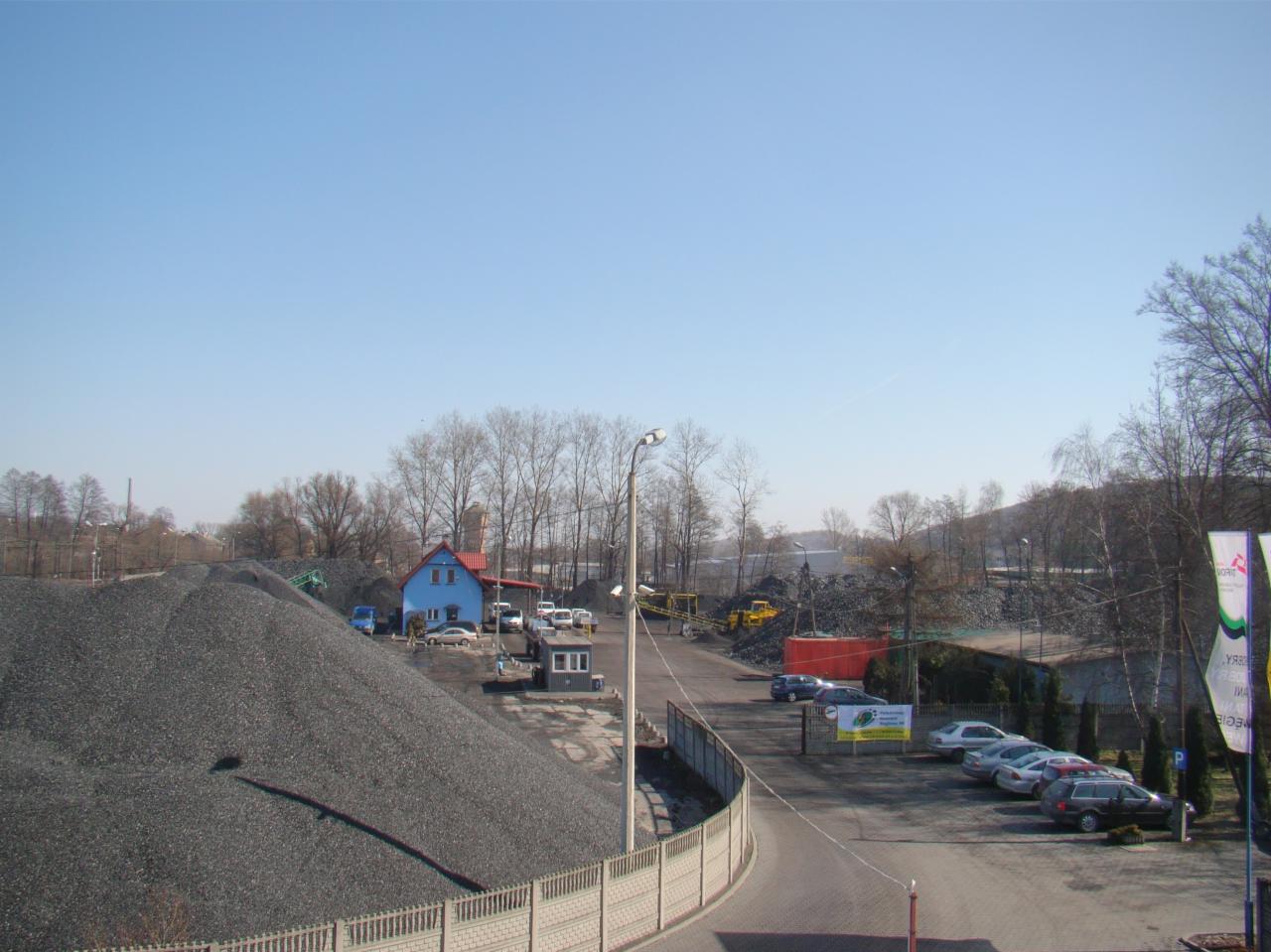 Widok z góry na plac składowania węgla; duży pryzmat czarnego węgla na pierwszym planie, w tle budynek biurowy, zaparkowane samochody i hałda węgla; żółta koparka w oddali.