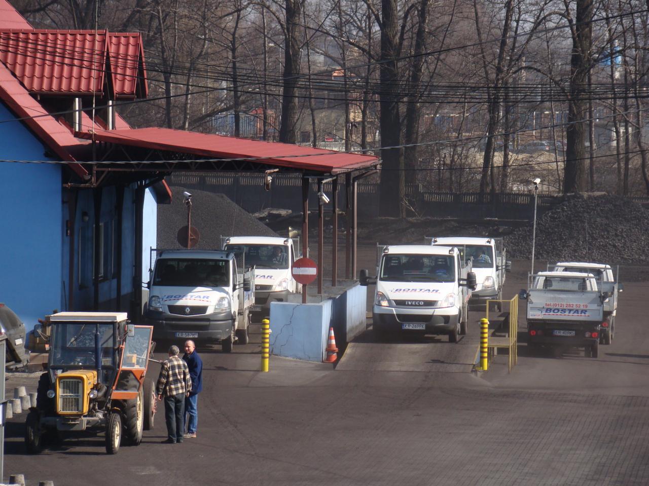 Teren składu opału z zaparkowanymi samochodami dostawczymi firmy transportowej, żółty traktor z otwartymi drzwiami i dwaj mężczyźni rozmawiający obok, w tle hałda węgla i budynek z czerwonym dachem.