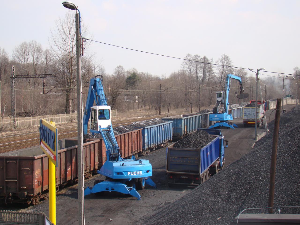 Niebieskie koparki Fuchs ładują węgiel z wagonów kolejowych do ciężarówek, widoczny znak informacyjny oraz tory kolejowe w tle.