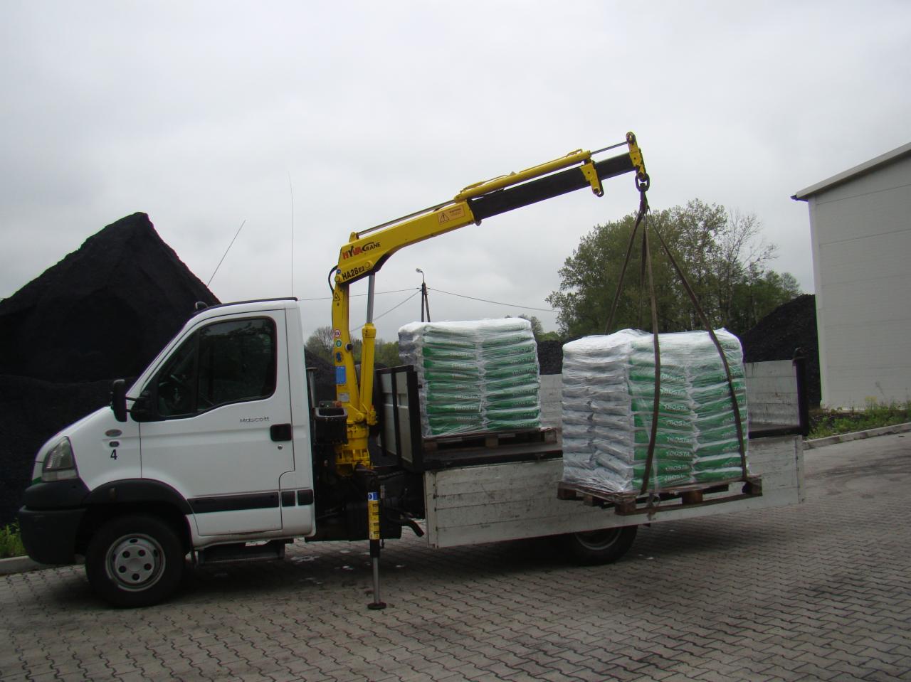 Biała ciężarówka z żółtym dźwigiem podnosi paletę z workami na tle hałdy węgla