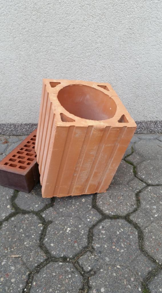 Dwa rodzaje cegieł: pustak kominowy z otworem i cegła dziurawka, leżące na szarym chodniku z kostki.