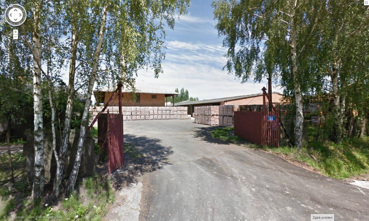 Otwarta, czerwona brama wjazdowa na teren z paletami cegieł, widok z drogi publicznej, w tle budynek magazynowy.