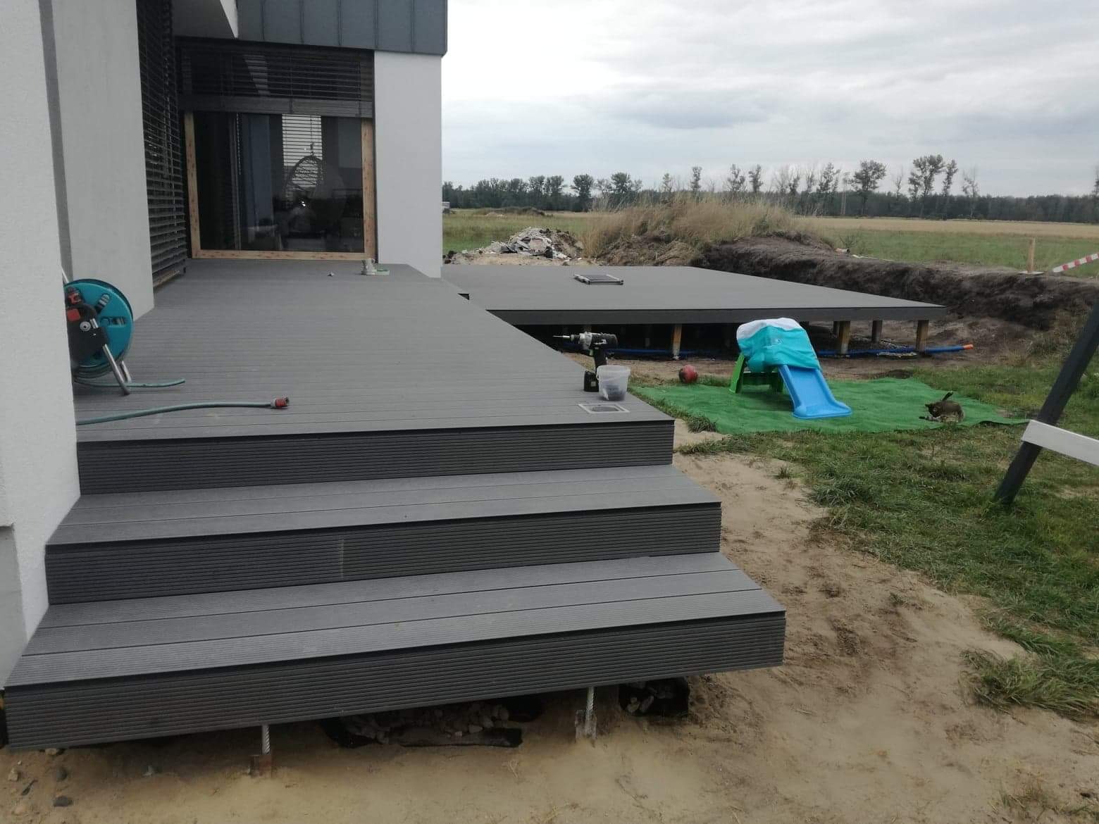 Montaż szarego tarasu z desek kompozytowych z widocznymi schodami i narzędziami, na tle placu budowy z zabawkami dla dzieci.