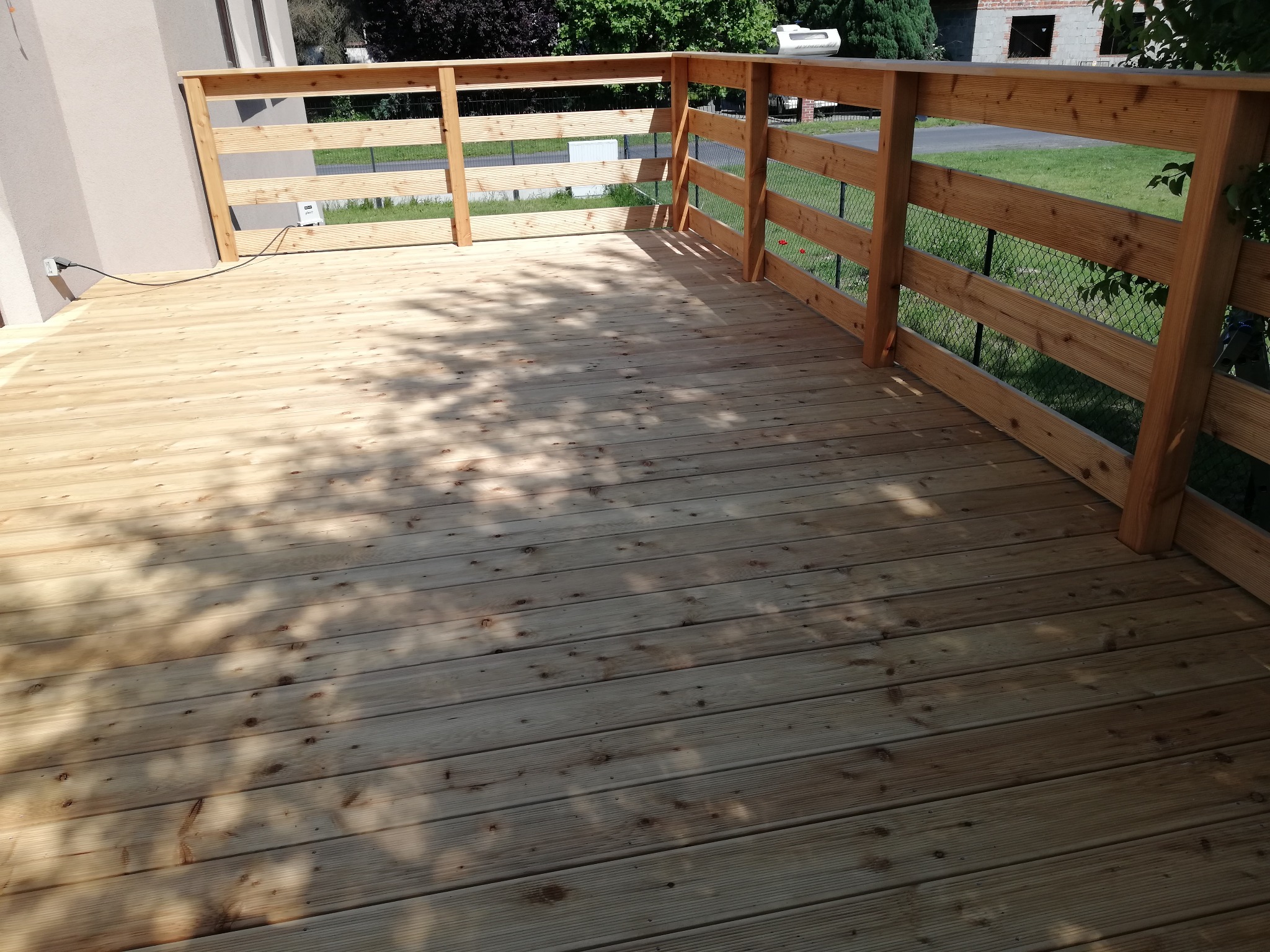 Świeżo położony taras z jasnego drewna z drewnianą balustradą, w tle zieleń i budynek z cegły.