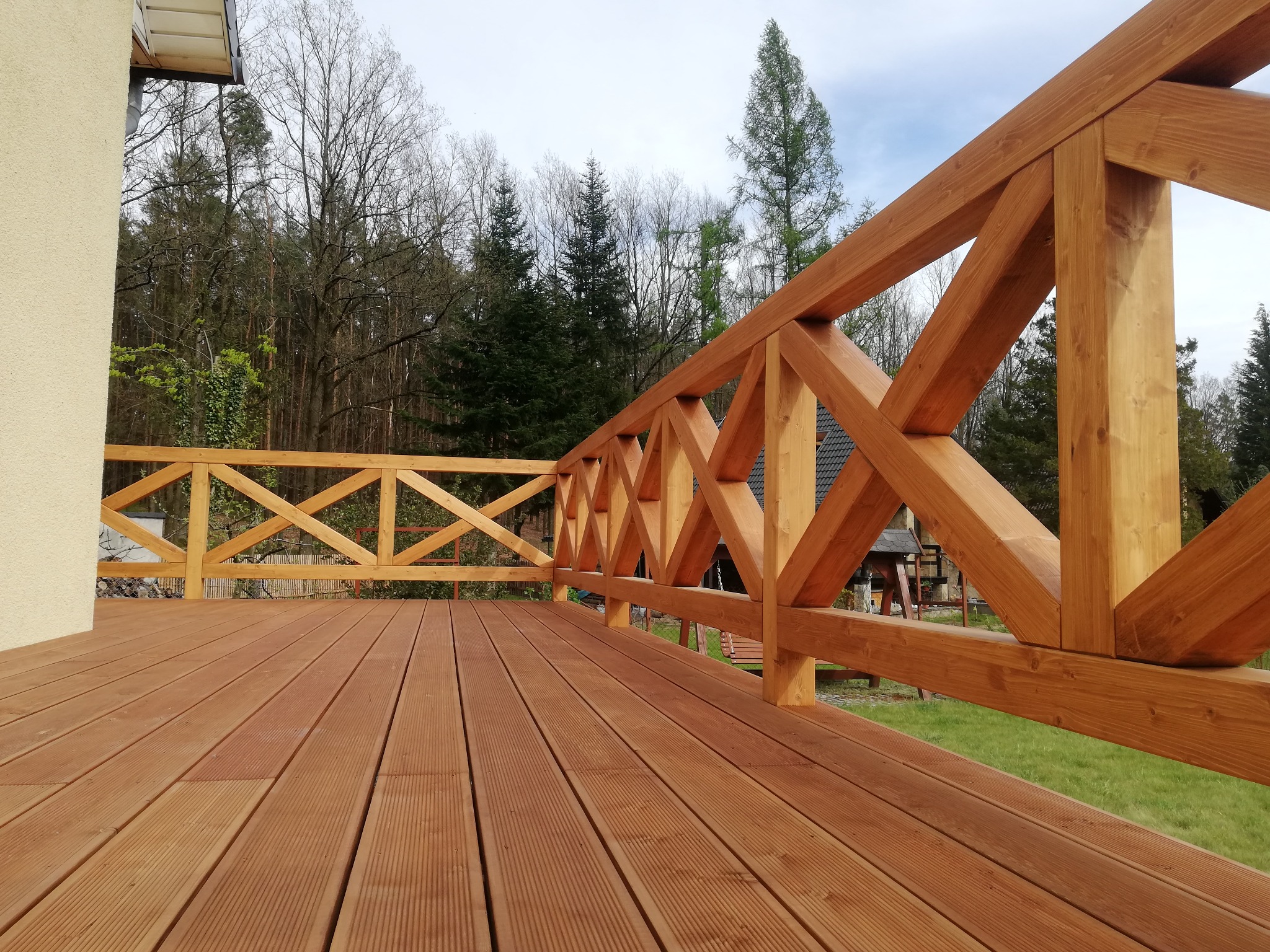 Nowy, drewniany taras z jasnymi deskami i balustradą w geometryczne wzory, widok na zewnątrz z zielenią i drzewami w tle.