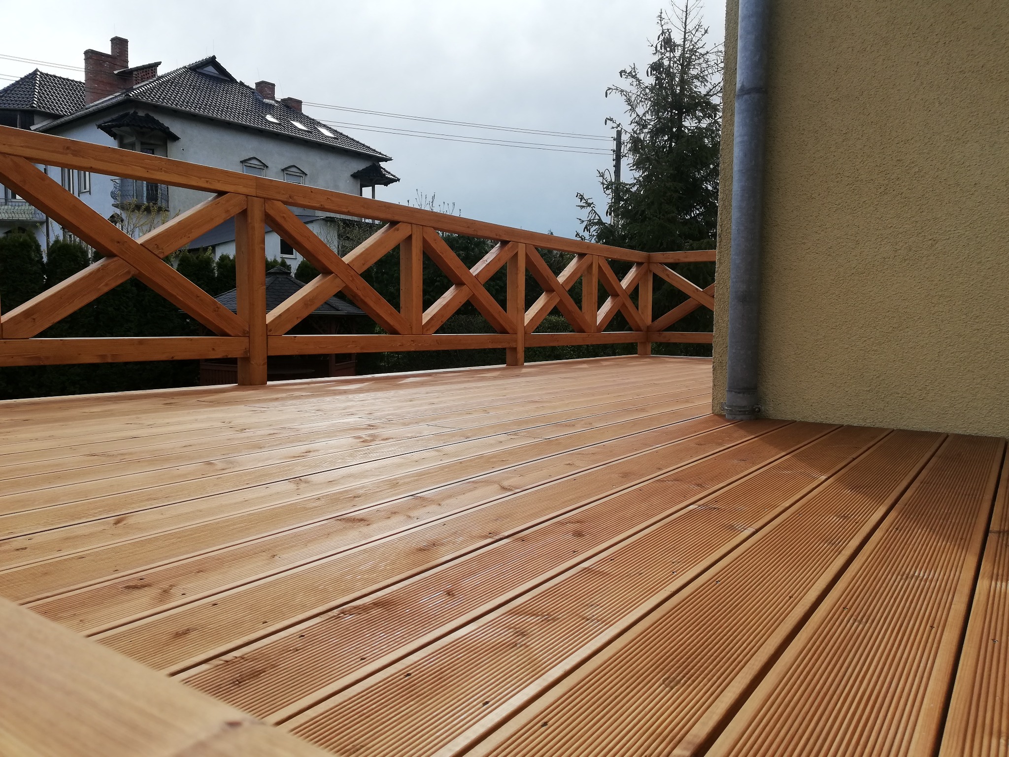 Nowo wybudowany taras z desek z drewnianą balustradą o wzorze krzyżowym, widok na dom sąsiada i rynnę odprowadzającą wodę deszczową.