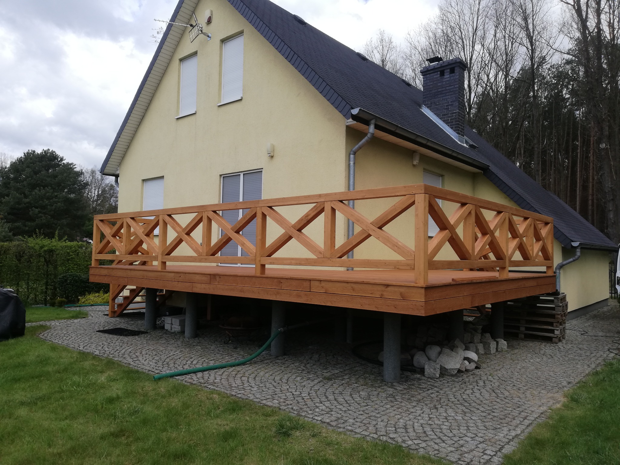 Nowoczesny taras z drewnianą balustradą w kształcie krzyży, wsparty na betonowych słupach, przylegający do żółtego domu z ciemną dachówką, widok z poziomu trawnika.