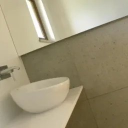 Nowoczesna łazienka z białą, owalną umywalką nablatową, minimalistyczną baterią podtynkową i szarą, kamienną okładziną ściany.