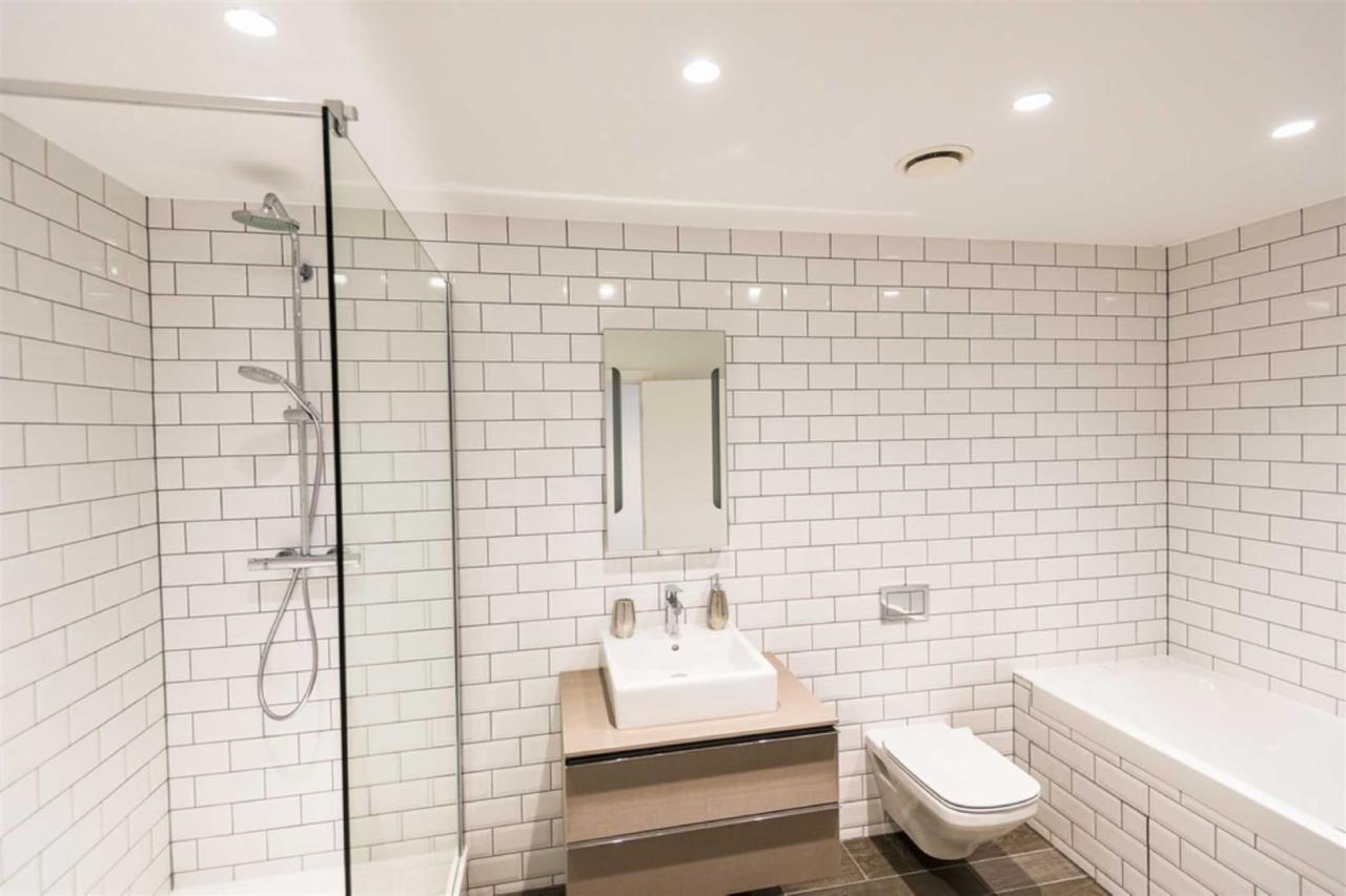 Nowoczesna łazienka z białymi, prostokątnymi płytkami, kabiną prysznicową ze szklaną ścianką, umywalką na szafce z drewnianym frontem i podwieszaną toaletą.