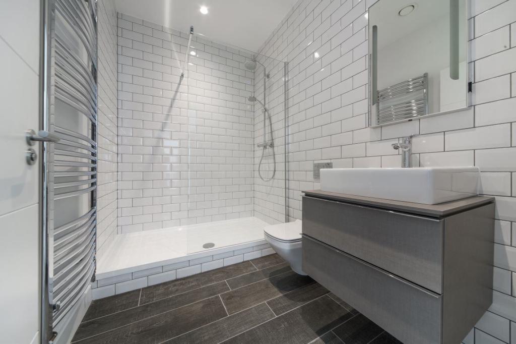 Nowoczesna łazienka z białymi, prostokątnymi płytkami ułożonymi w cegiełkę, szarą szafką pod umywalką, kabiną prysznicową typu walk-in i grzejnikiem drabinkowym po lewej stronie.
