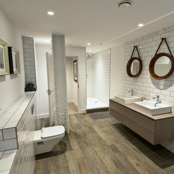 Przestronna łazienka w stylu industrialnym z dwoma umywalkami nablatowymi, kabiną prysznicową typu walk-in i podłogą imitującą drewno. Charakterystyczna kolumna konstrukcyjna w centrum pomieszczenia.