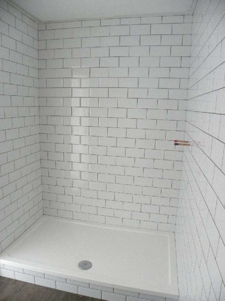 Biały brodzik z odpływem i ścianami wyłożonymi białymi, prostokątnymi płytkami z ciemną fugą, widoczne wystające rurki instalacji wodnej.