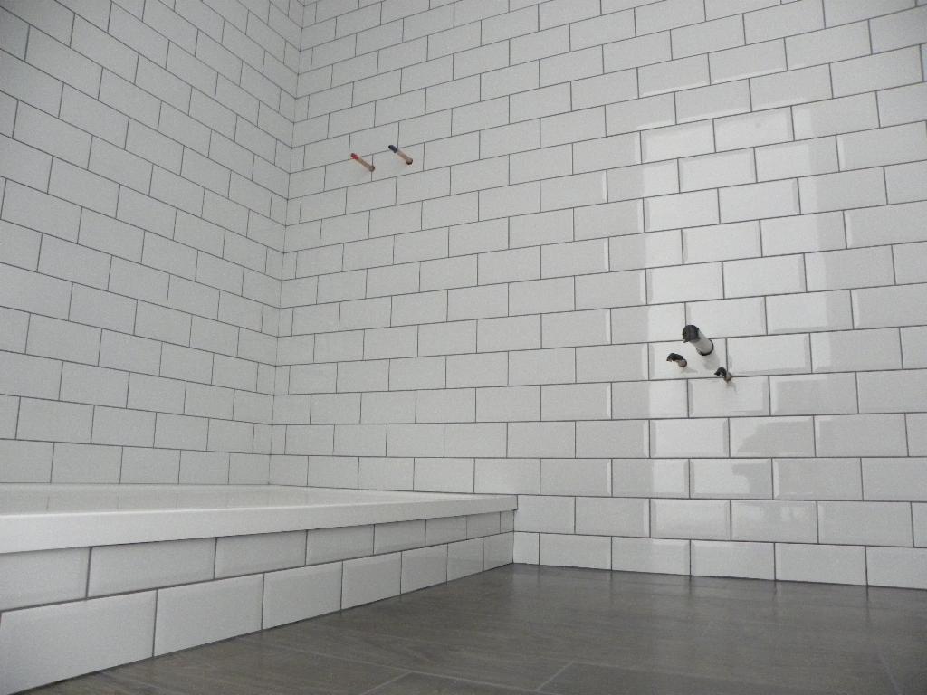 Ściana łazienki wyłożona białymi, błyszczącymi płytkami typu metro z szarą fugą, widoczne wystające rury instalacji wodnej i kanalizacyjnej, oraz fragment obudowy wanny.