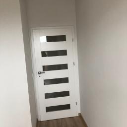 Budgro - Nowoczesne białe drzwi wewnętrzne z poziomymi przeszkleniami w wąskim korytarzu z panelami podłogowymi.