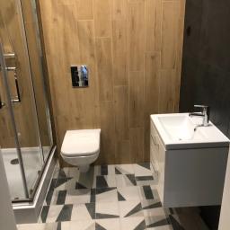 Budgro - Wykończona łazienka z drewnopodobnymi płytkami na ścianach, kabiną prysznicową, podwieszaną toaletą i umywalką z szafką, na podłodze geometryczne płytki w odcieniach szarości.