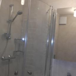 Budgro - Szklana kabina prysznicowa z chromowanymi detalami, zamontowana w narożniku wyłożonym beżowymi płytkami, widok na umywalkę i lustro z odbiciem wentylatora.