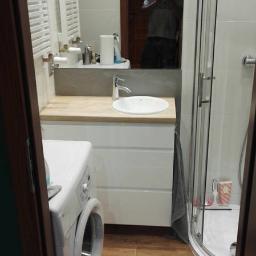 Budgro - Ukończona łazienka z drewnopodobną podłogą, białą szafką pod umywalką z drewnianym blatem, pralką z przodu i kabiną prysznicową z zaokrąglonym narożnikiem; w lustrze odbija się osoba robiąca zdjęcie.