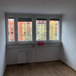 Pusty pokój z panelami podłogowymi, grzejnikiem i oknem z widokiem na kolorowy blok mieszkalny; na parapecie leżą różowe rękawice robocze.