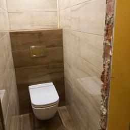 Budgro - Toaleta w trakcie montażu we Wrocławiu, widoczne płytki imitujące marmur na ścianach i drewno na podłodze, ściana z cegły po prawej stronie w trakcie wykończenia.