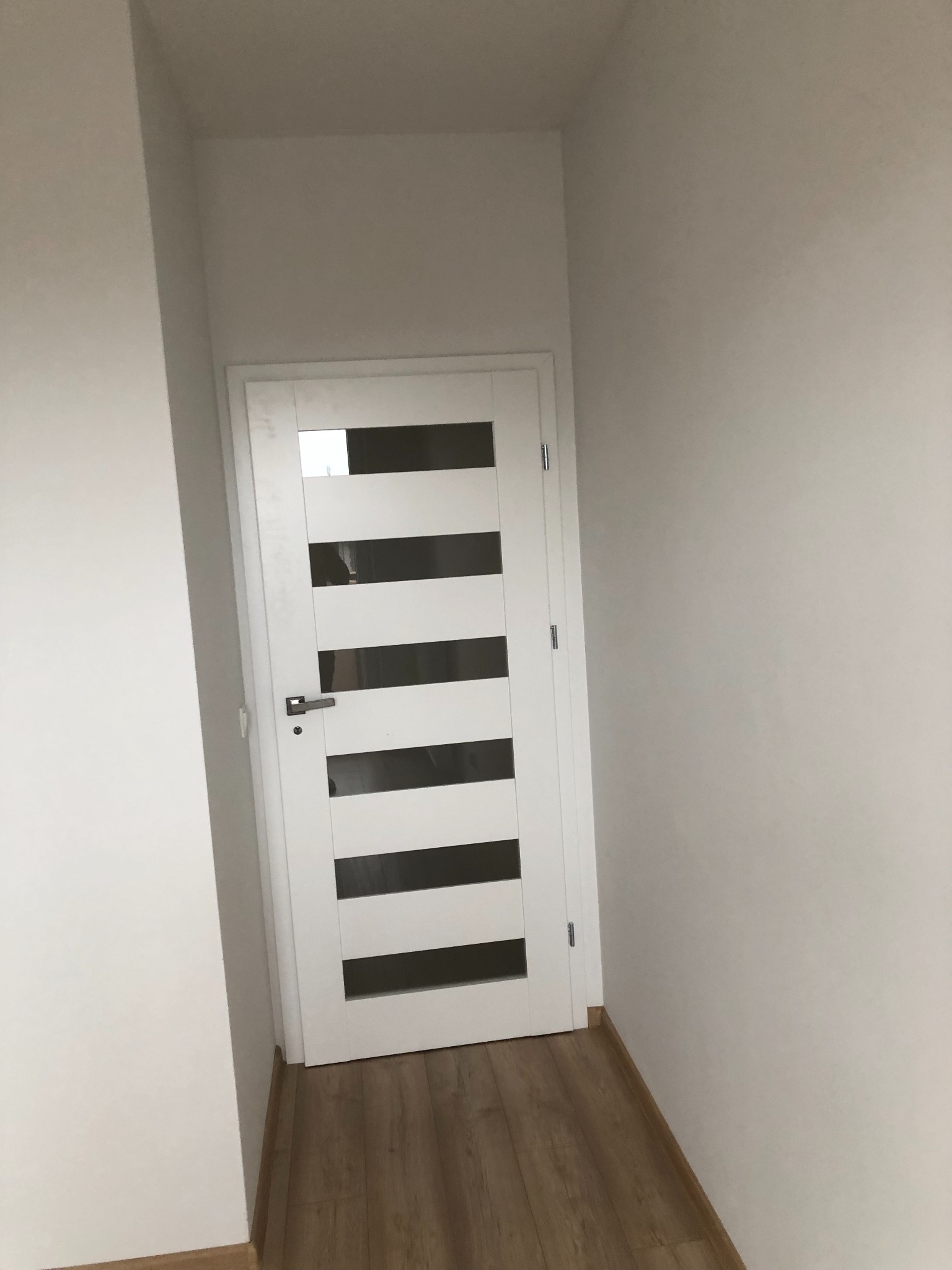 Nowoczesne białe drzwi wewnętrzne z poziomymi przeszkleniami w wąskim korytarzu z panelami podłogowymi.