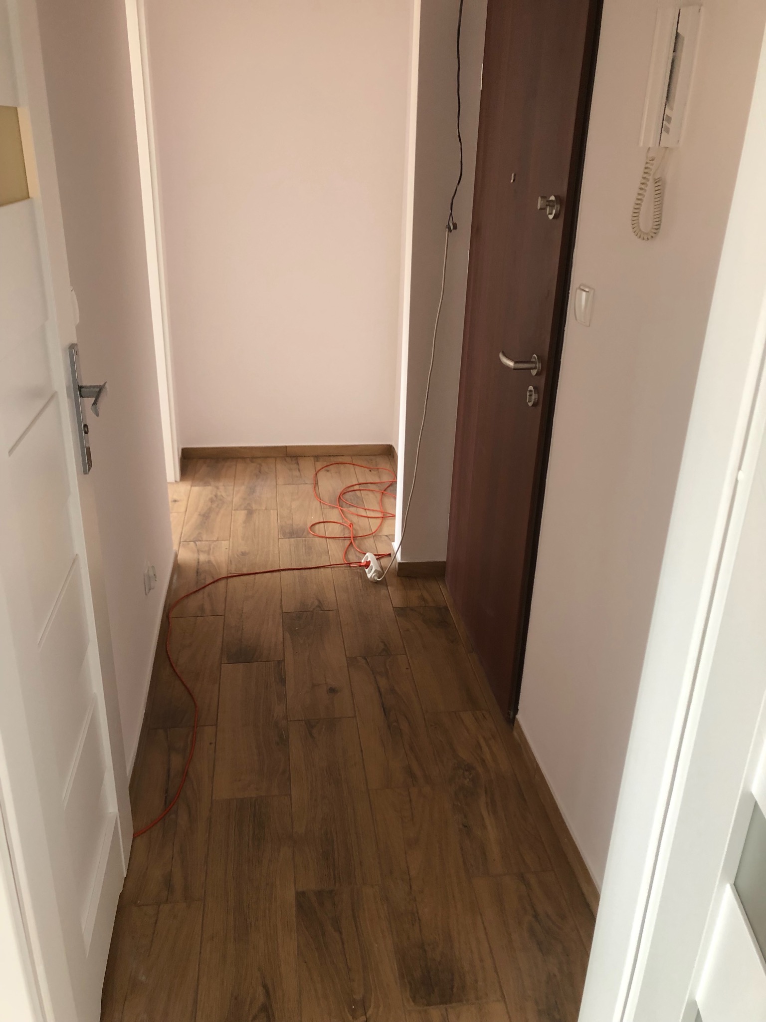 Korytarz po ułożeniu drewnopodobnych paneli podłogowych, widoczny kabel elektryczny i gniazdko, białe ściany i drzwi.