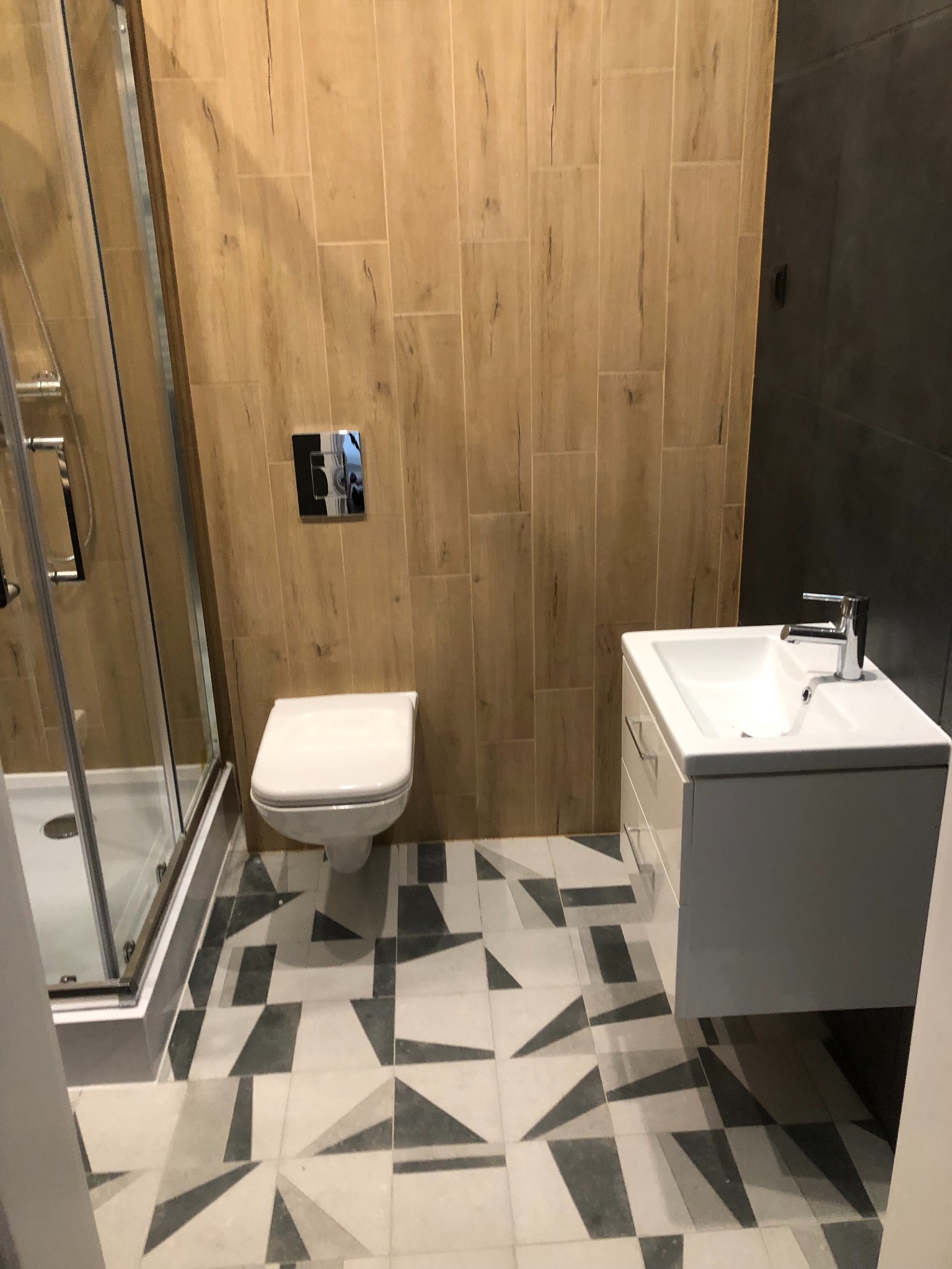 Wykończona łazienka z drewnopodobnymi płytkami na ścianach, kabiną prysznicową, podwieszaną toaletą i umywalką z szafką, na podłodze geometryczne płytki w odcieniach szarości.