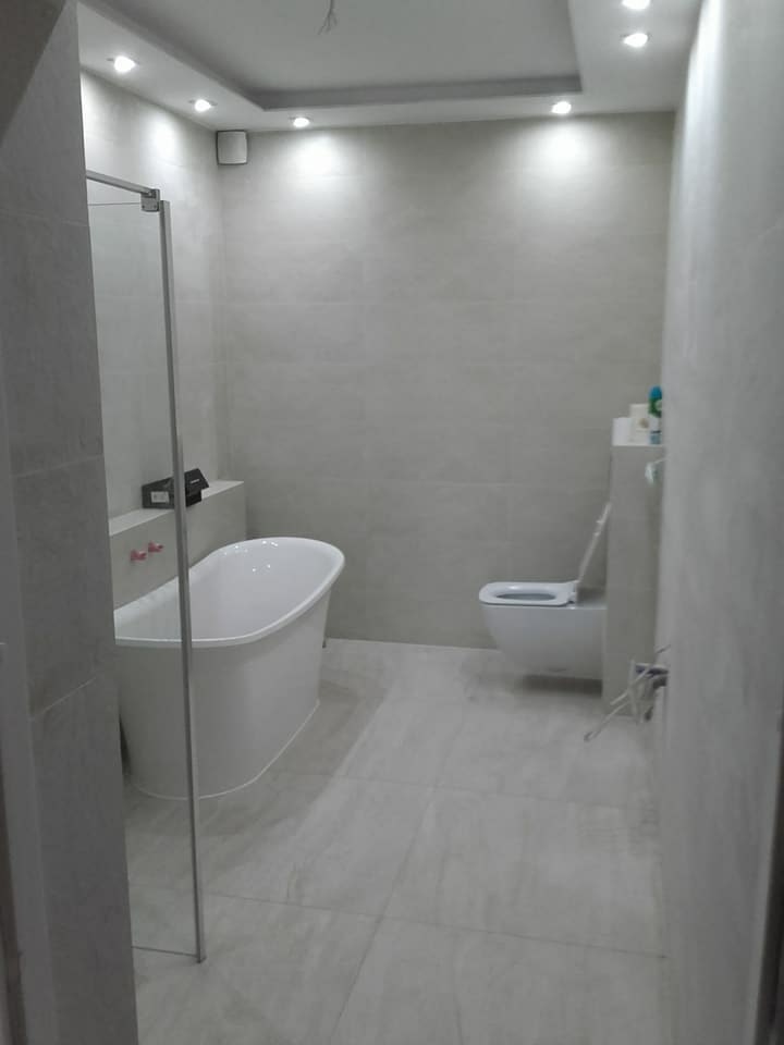 Nowoczesna łazienka w odcieniach szarości z wolnostojącą wanną, toaletą w zabudowie i szklanym panelem prysznicowym, wykończona płytkami imitującymi kamień