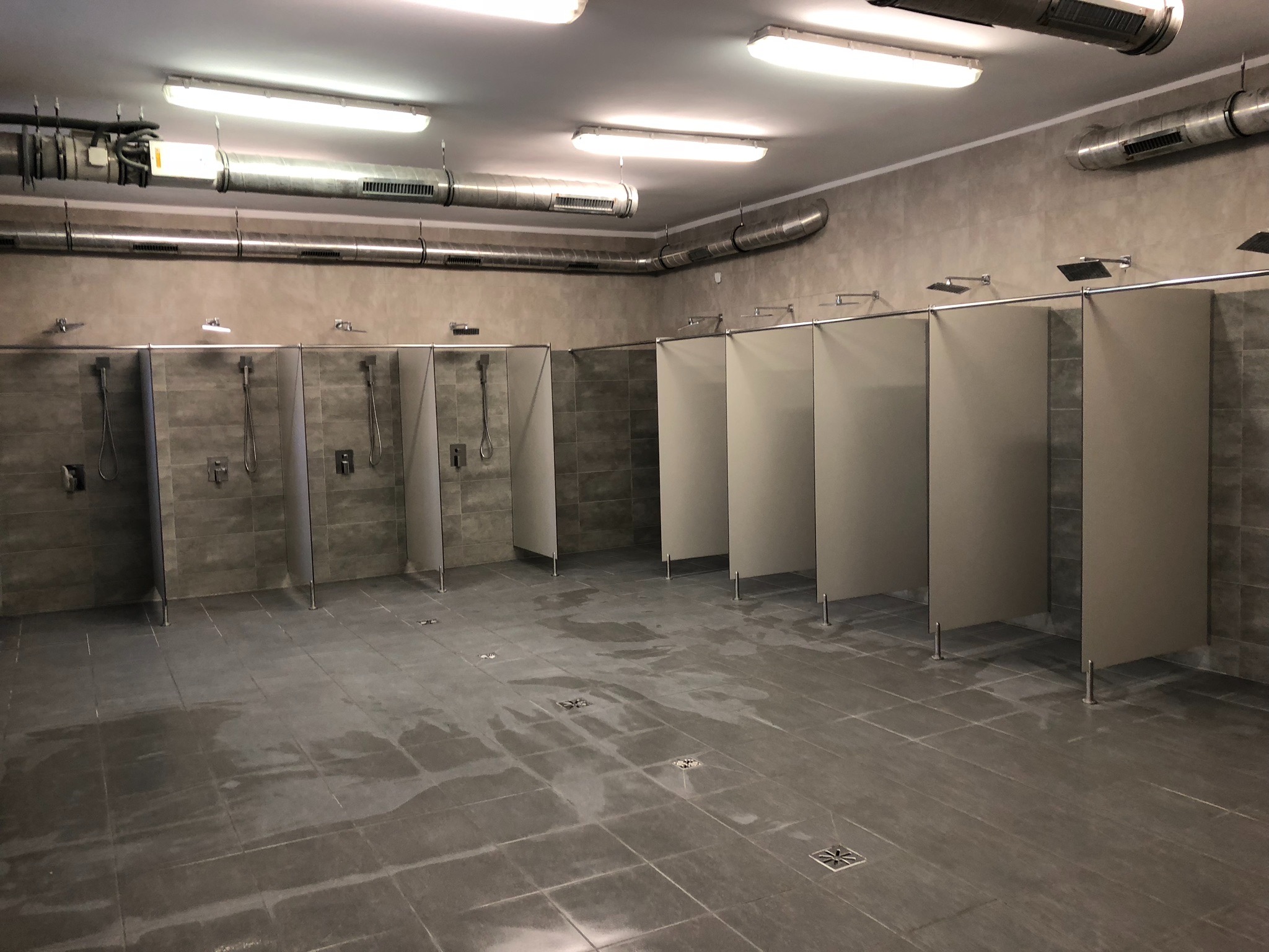 Wnętrze szarej, publicznej łazienki z kabinami prysznicowymi i widocznymi instalacjami wentylacyjnymi pod sufitem.