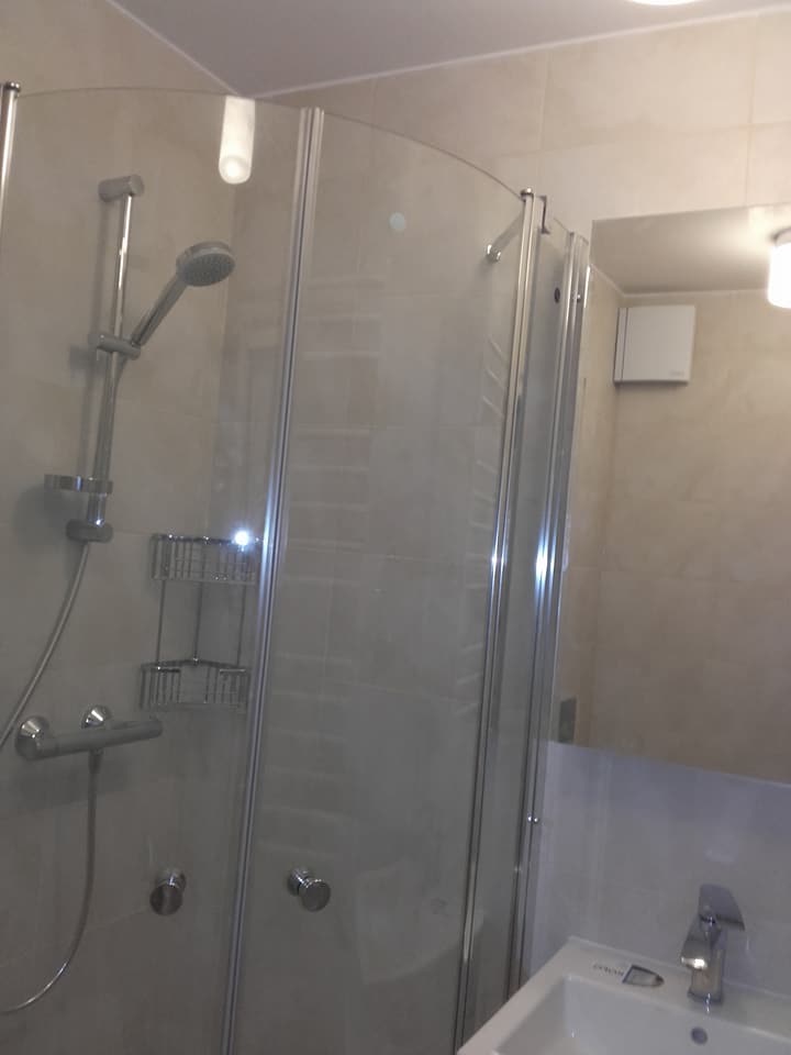 Szklana kabina prysznicowa z chromowanymi detalami, zamontowana w narożniku wyłożonym beżowymi płytkami, widok na umywalkę i lustro z odbiciem wentylatora.