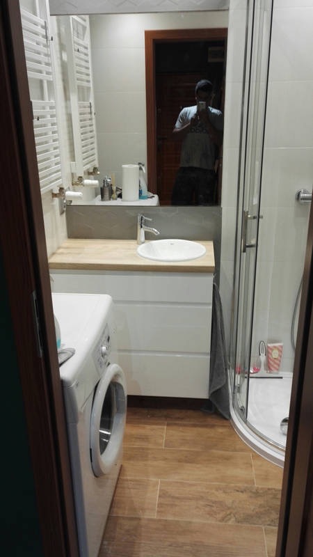 Ukończona łazienka z drewnopodobną podłogą, białą szafką pod umywalką z drewnianym blatem, pralką z przodu i kabiną prysznicową z zaokrąglonym narożnikiem; w lustrze odbija się osoba robiąca zdjęcie.