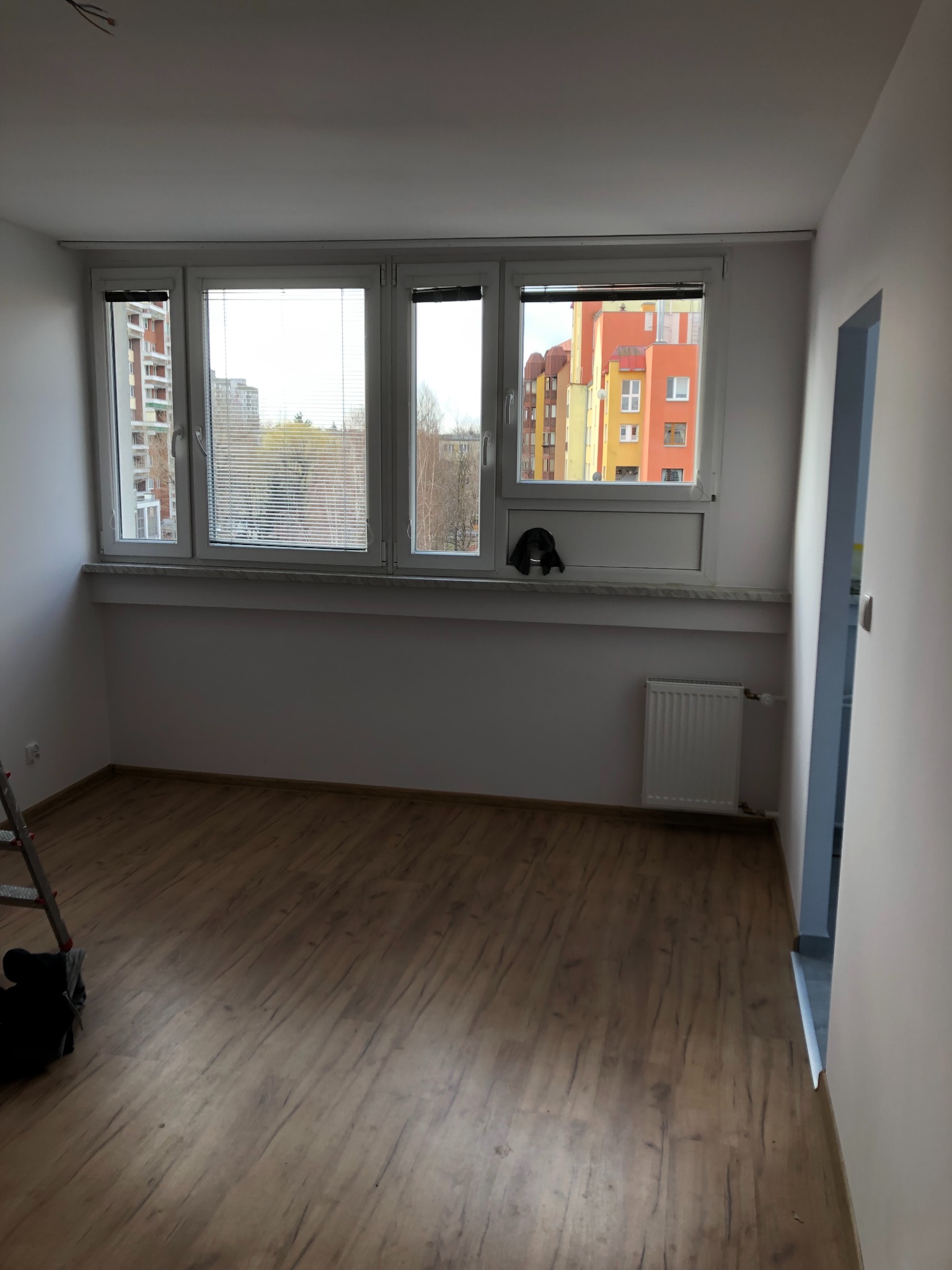 Pusty pokój z drewnianą podłogą, białymi ścianami i trzema oknami z żaluzjami, widok na bloki mieszkalne za oknem, drabina oparta o ścianę w lewym dolnym rogu.