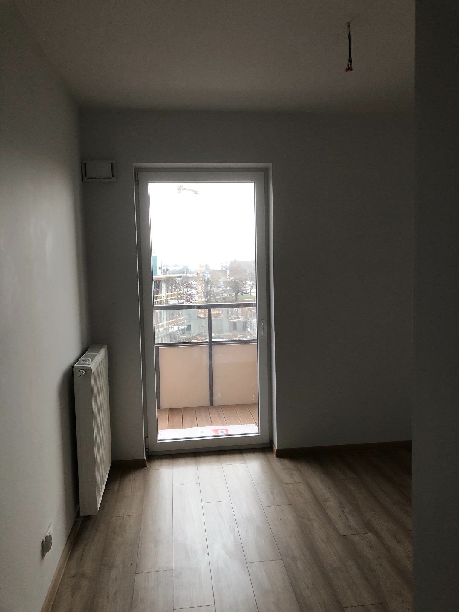 Pusty pokój z panelami podłogowymi, grzejnikiem i balkonem, widok na plac budowy za oknem.