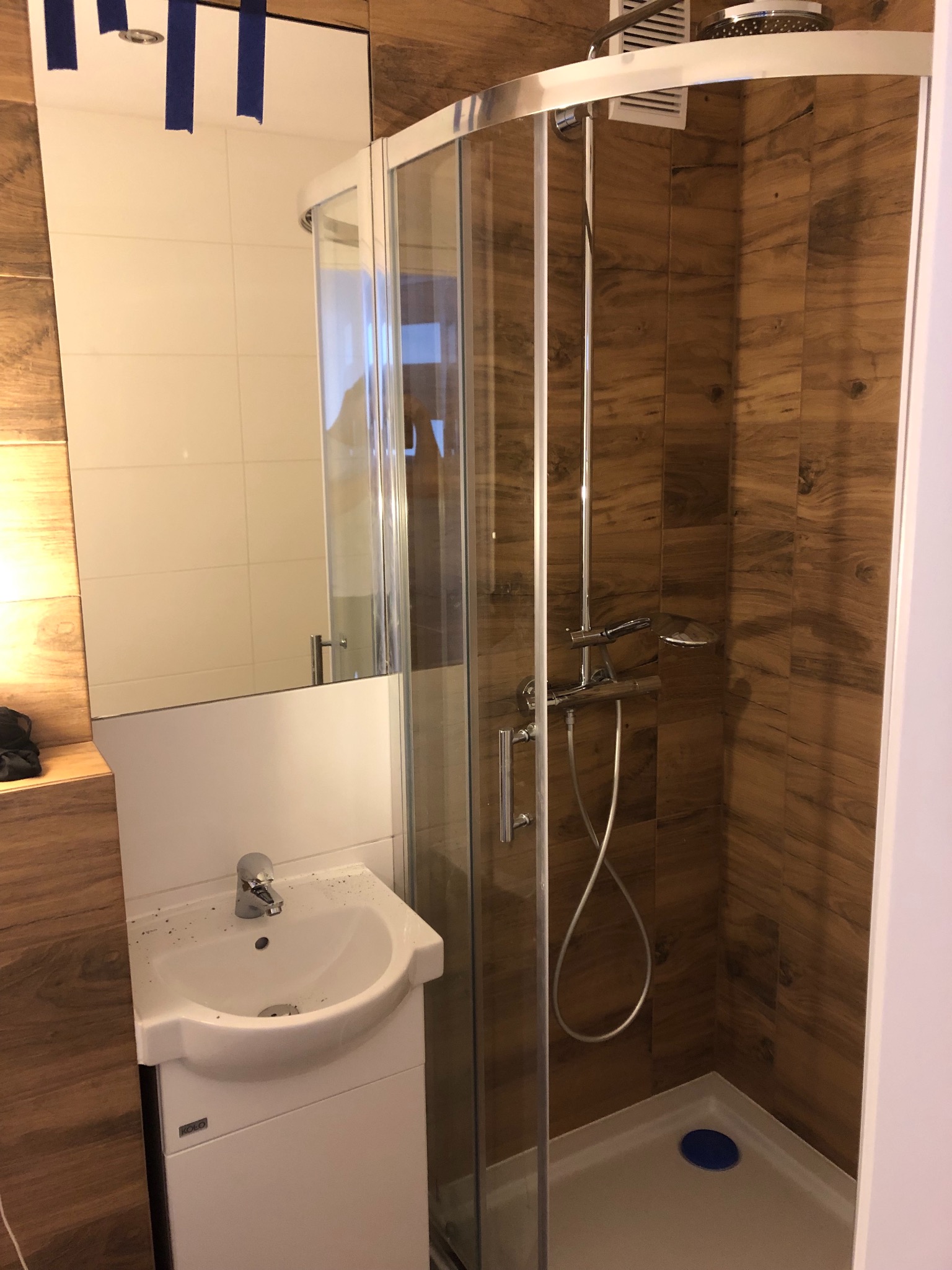 Kabina prysznicowa z drewnopodobnymi płytkami, umywalka KOLO z szafką i lustro zabezpieczone taśmą malarską w łazience.