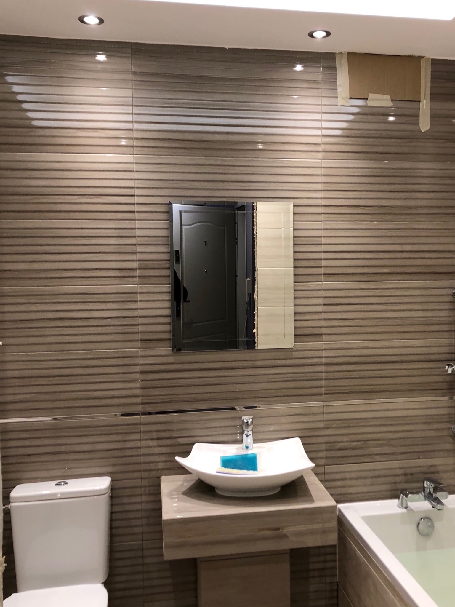 Nowoczesna łazienka z umywalką w kształcie łódki, toaletą kompaktową i wanną, wyłożona poziomymi płytkami imitującymi drewno, lustro z widocznym odbiciem drzwi wejściowych, zabezpieczone kartonem...