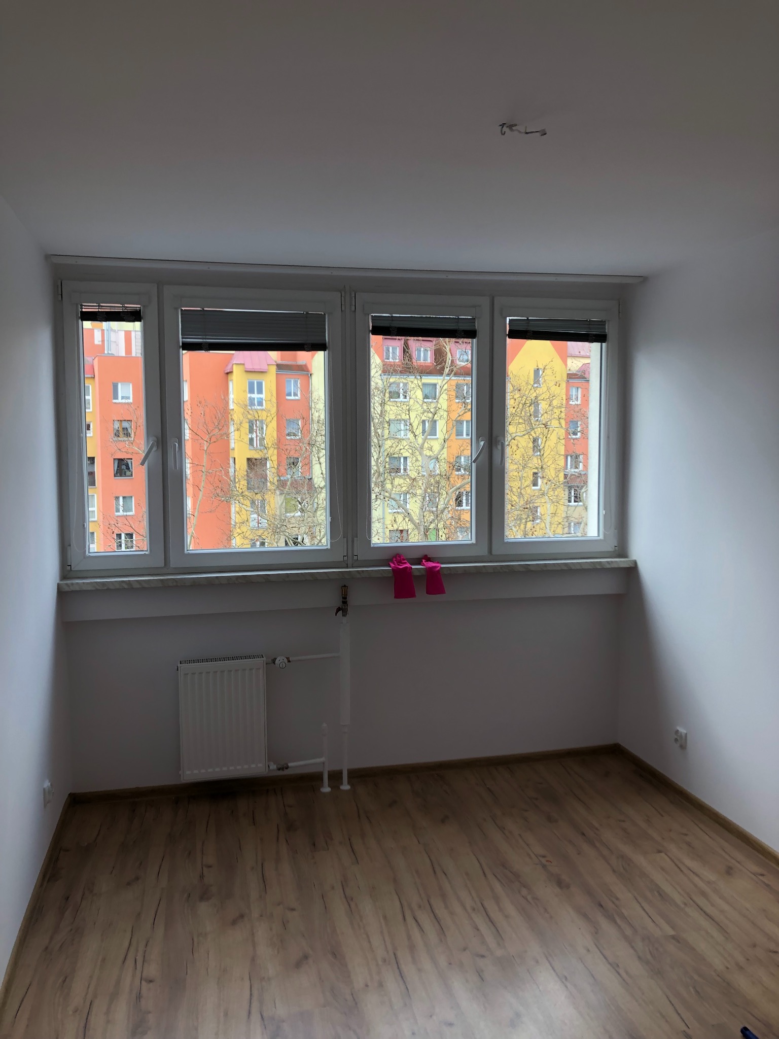 Pusty pokój z panelami podłogowymi, grzejnikiem i oknem z widokiem na kolorowy blok mieszkalny; na parapecie leżą różowe rękawice robocze.