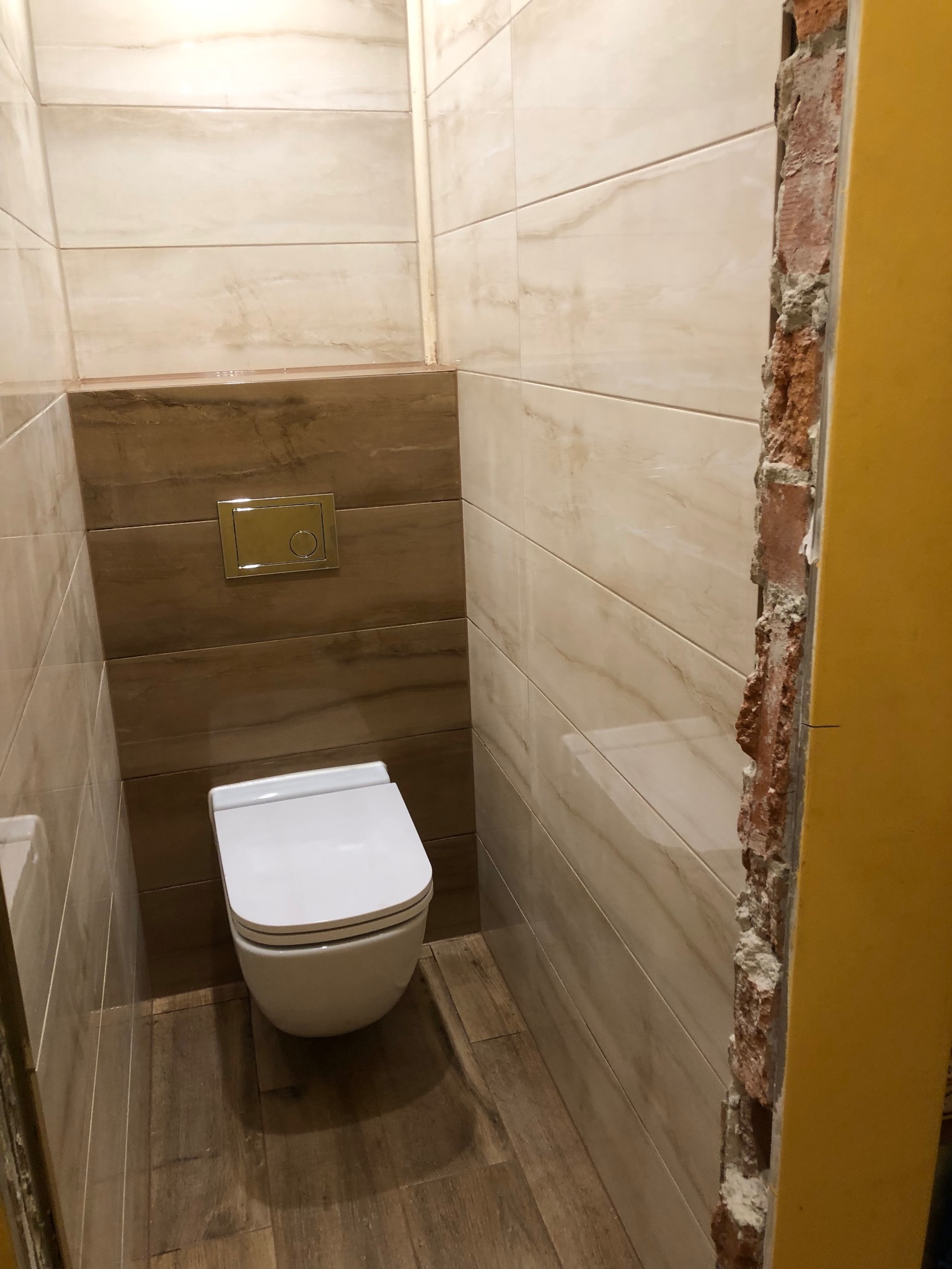 Toaleta w trakcie montażu we Wrocławiu, widoczne płytki imitujące marmur na ścianach i drewno na podłodze, ściana z cegły po prawej stronie w trakcie wykończenia.