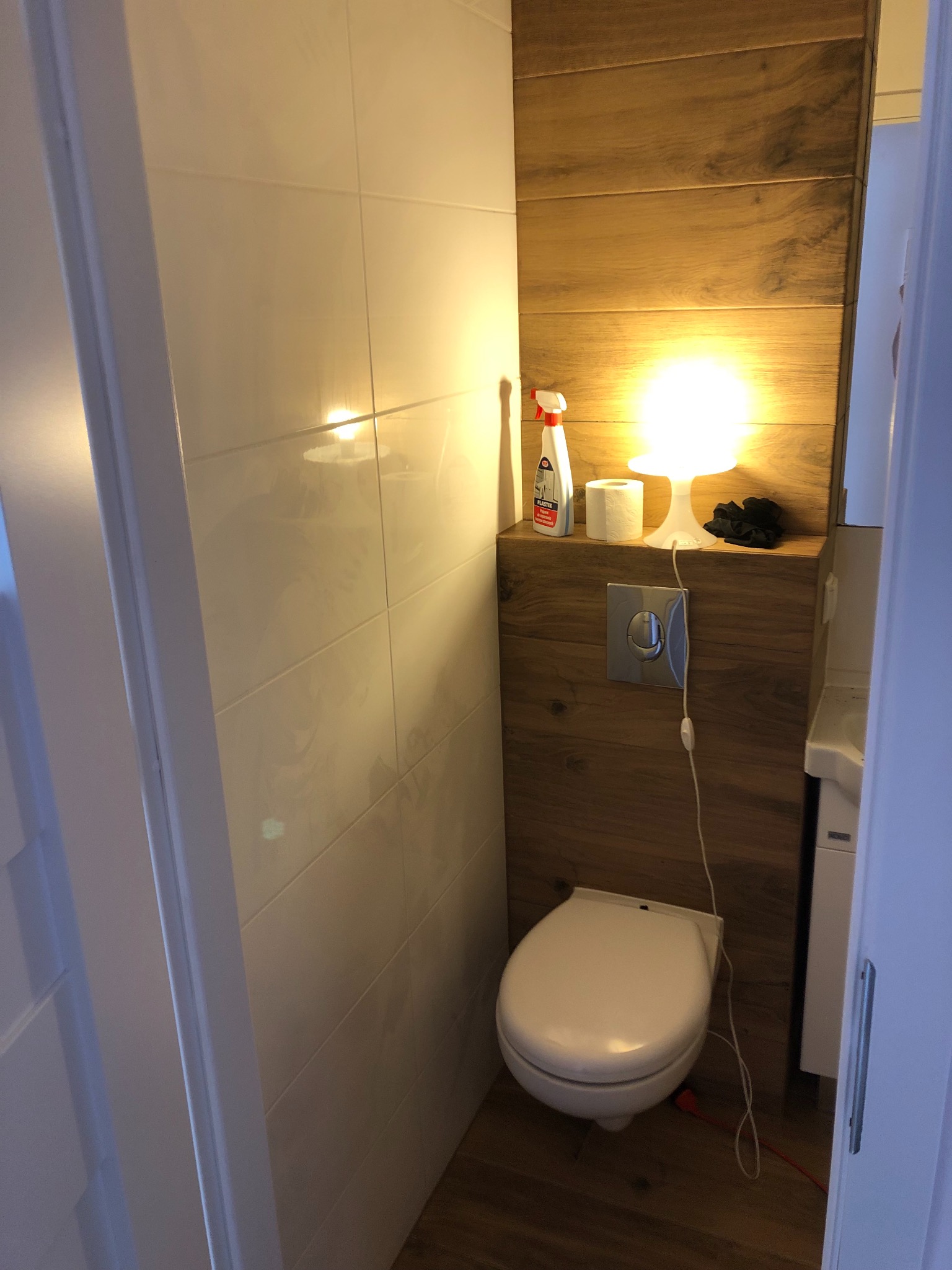 Nowoczesna toaleta z drewnianą ścianą, białymi płytkami i oświetleniem punktowym, widoczny dozownik mydła i rolka papieru toaletowego