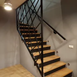 Nowoczesne schody z drewnianymi stopniami i czarną, geometryczną balustradą z metalu, oświetlone wiszącą lampą.