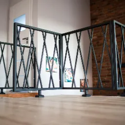 Nowoczesna, czarna balustrada z metalu z motywem krzyżujących się ukośnych prętów, zamontowana na antresoli w domu z ceglaną ścianą w tle i zdjęciami rodzinnymi na ścianie.