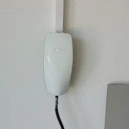 Biała stacja ładowania Tesla Wall Connector zamontowana na ścianie, z widocznym przewodem zasilającym zabezpieczonym opaską zaciskową.