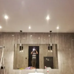 Nowoczesna łazienka z szarą marmurową okładziną, oświetleniem punktowym i wiszącymi lampami. W odbiciu lustrzanym widoczna osoba robiąca zdjęcie. Rozstawiona drabina sugeruje prace instalacyjne...