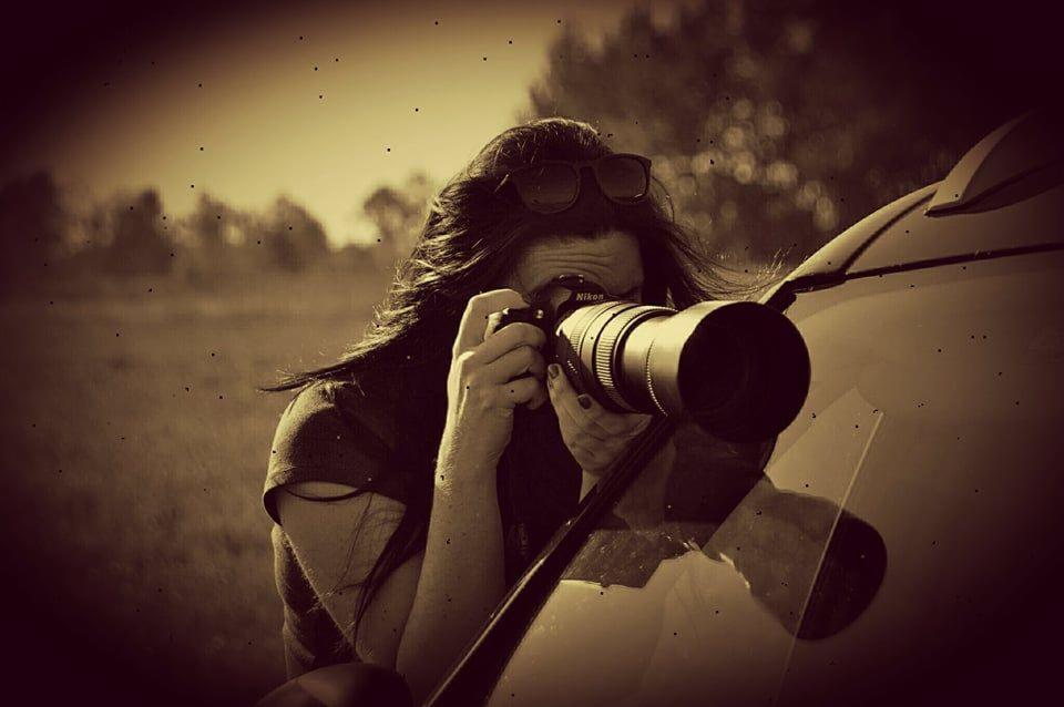 Kobieta z długimi ciemnymi włosami robi zdjęcie profesjonalnym aparatem Nikon z dużym obiektywem, stojąc obok błyszczącego samochodu na tle łąki i drzew, w sepiowej tonacji.
