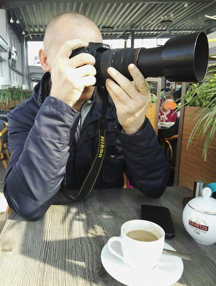 Mężczyzna z ogoloną głową, ubrany w granatową kurtkę, trzyma aparat Nikon z założonym teleobiektywem, siedząc przy drewnianym stole z filiżanką kawy, telefonem i cukiernicą Basilur.