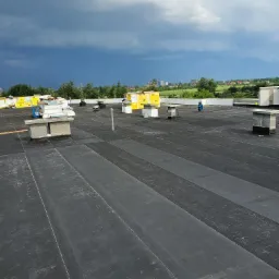 Płaski dach pokryty czarną papą termozgrzewalną, widoczne kominki wentylacyjne i materiały izolacyjne przygotowane do montażu, w tle burzowe chmury nad zielonym krajobrazem.