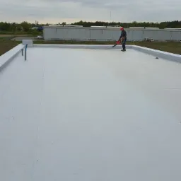 Pracownik w czerwonej czapce używa dmuchawy na płaskim, białym dachu budynku przemysłowego, widoczne orynnowanie i elementy instalacji. W tle zabudowania przemysłowe i drzewa pod pochmurnym niebem.