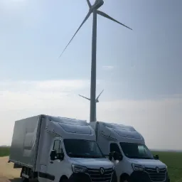Dwa białe busy Renault Master Longcab zaparkowane na polnej drodze z turbiną wiatrową w tle, w słoneczny dzień.