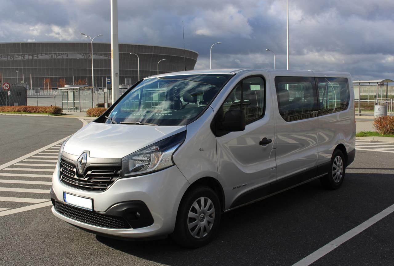 Srebrny bus Renault Trafic na parkingu z widokiem na stadion w tle, zaparkowany na pasach dla pieszych.